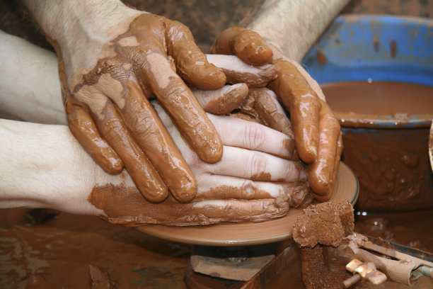 一个工匠的手引导着另一只手，教他如何用陶瓷轮工作。图片下载