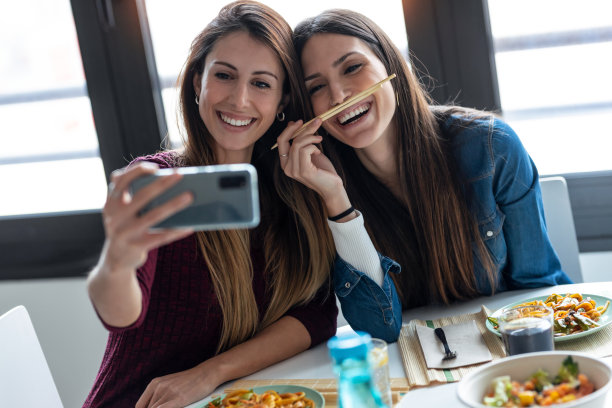 两个微笑美丽的女性朋友吃日本食物，同时在家里的厨房用智能手机自拍。图片下载