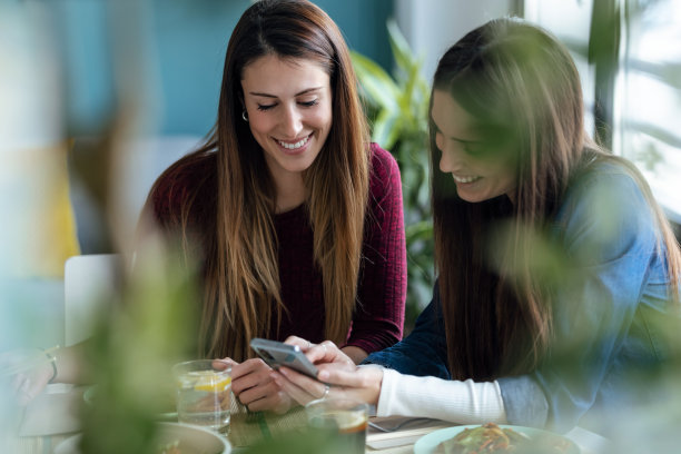 两个漂亮的女人朋友吃日本食物，同时在厨房看智能手机在家。图片下载
