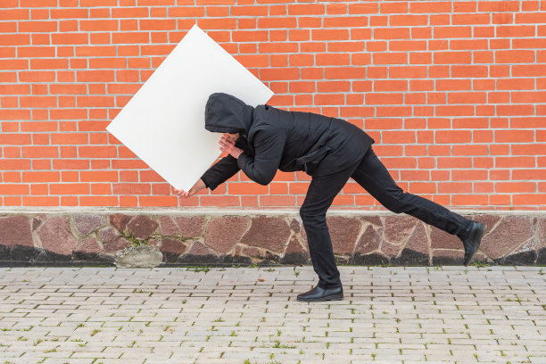一名男子拿着一张空白的白色画布，弯曲着身体沿着一堵红砖墙移动图片下载