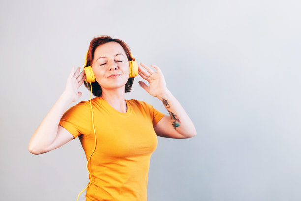 穿着亮t恤，戴着黄色耳机的漂亮女人。灰色的墙壁与复印空间。图片下载