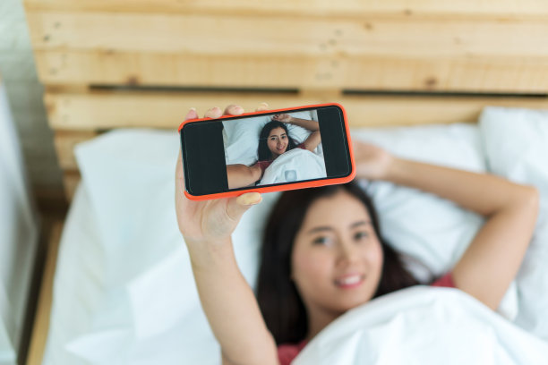 漂亮快乐的年轻女子在白色的床上用智能手机自拍。对焦手握移动。图片下载