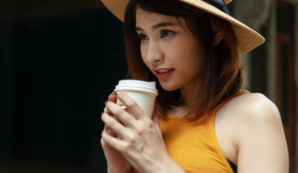 有吸引力的女人站在混凝土墙上拿着咖啡杯的背景与复制空间。华丽的女人穿着休闲的衣服和帽子喜欢喝咖啡，使她新鲜。图片下载