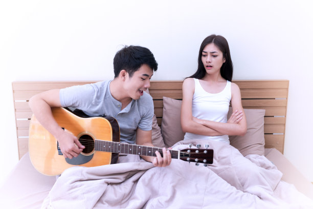 不快乐的年轻女子在弹原声吉他时惹恼了一个男人。美丽的女孩感觉不满她的男朋友在卧室大声唱歌。夫妻关系有问题的概念。图片下载
