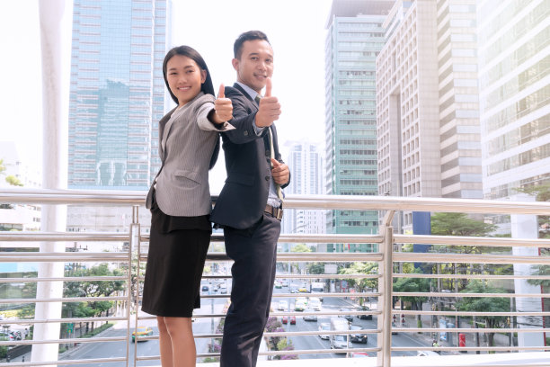 微笑的商业夫妇穿着西装，在一起竖起大拇指代表商业上的成功，好的，很好，所有的好东西，就像在现代城市里一样。成功的商业和团队合作理念。图片下载