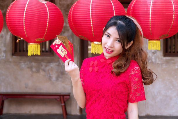 漂亮快乐的年轻女子穿着旗袍，手里拿着红色和金色的信封，里面装着好运和繁荣的钱，家人给她看相机。庆祝中国新年的概念图片下载