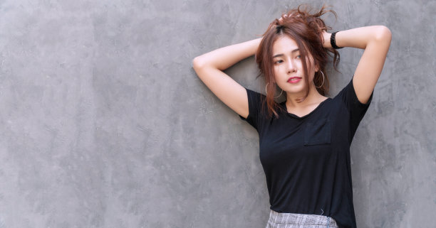 迷人的年轻女子感到放松抱着她的浓密的头发在头上灰色的墙壁背景。开朗的女孩把她的棕色波浪长发扎成马尾辫，看着有拷贝空间的东西。图片下载