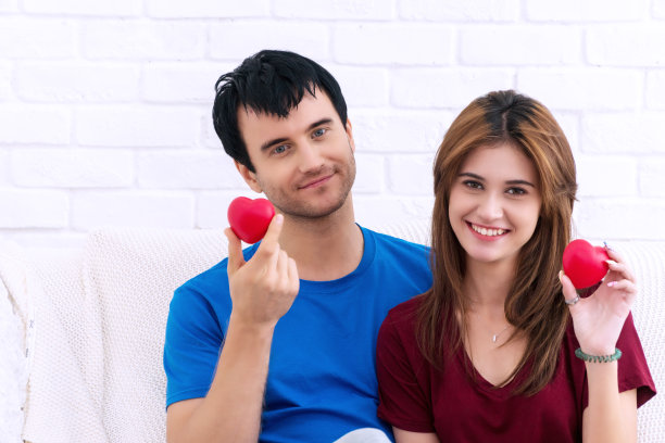 情人节。爱和关系的概念。喜气洋洋的年轻夫妇坐在白色的沙发上，手里捧着红心。图片下载