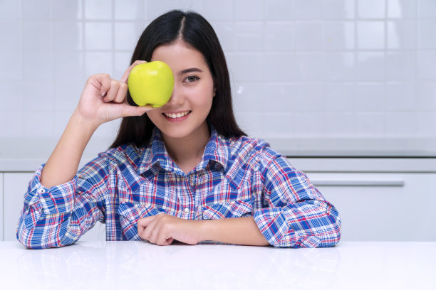 健康饮食的概念。迷人的微笑年轻女子拿着新鲜的绿色苹果盖住她的眼睛在她的厨房看起来的相机早餐。图片下载