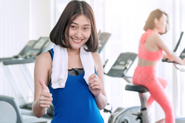 活跃的运动迷人的年轻愉快的微笑女人穿着运动服与毛巾举着她的拇指在健身中心锻炼后。健身苗条的女性站在健身房。运动的概念。图片下载