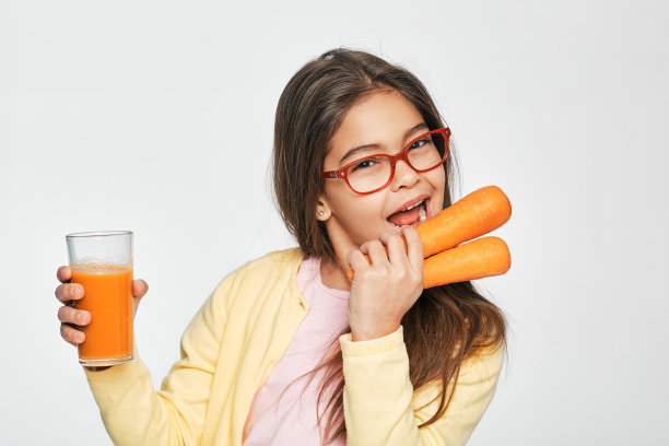 混血女孩吃胡萝卜和喝胡萝卜汁，在灰色背景。儿童的健康生活方式图片下载