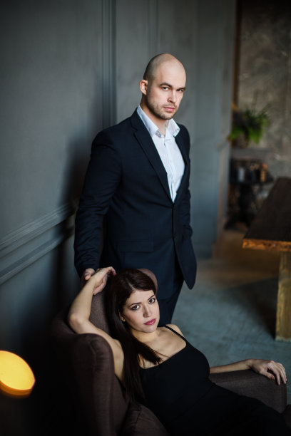优雅的情侣相爱。一男一女在暗室里。一个穿黑裙子的女孩坐在椅子上，一个穿西装的秃顶男人靠墙站在阁楼上。垂直的照片。情人节图片下载