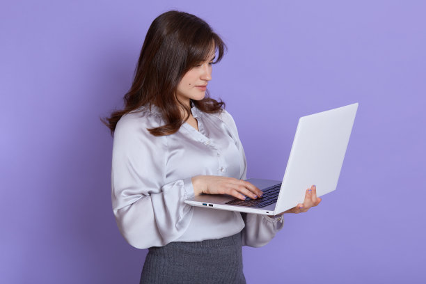 白种人美丽的年轻女子与手提电脑摆姿势靠在淡紫色的墙壁，看着设备屏幕，被集中而工作，穿着优雅的服装。图片下载