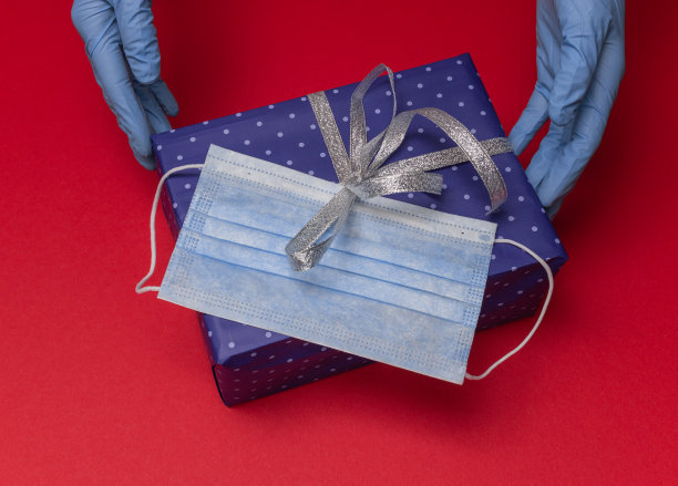 双手在蓝色乳胶手套持有蓝色礼盒在纸与圆点与丝带作为圣诞，新年，情人节的礼物在红色背景。最上面是医用口罩俯视图图片下载