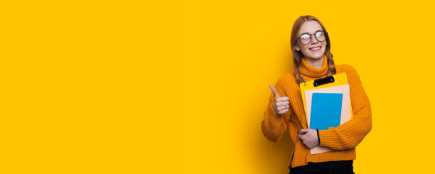 一个可爱的学生在工作室的黄色墙壁上指着类似的标志，在自由空间附近对着相机微笑，拿着文件夹图片下载