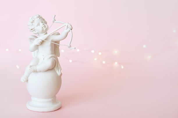 粉红色背景上带着蝴蝶结的天使丘比特小雕像。情人节图片下载