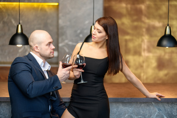 一对美丽情侣的约会。一男一女端着红酒站在吧台边。一个穿黑色开叉裙子的女孩和一个穿西装的秃顶男人。情人节。图片下载