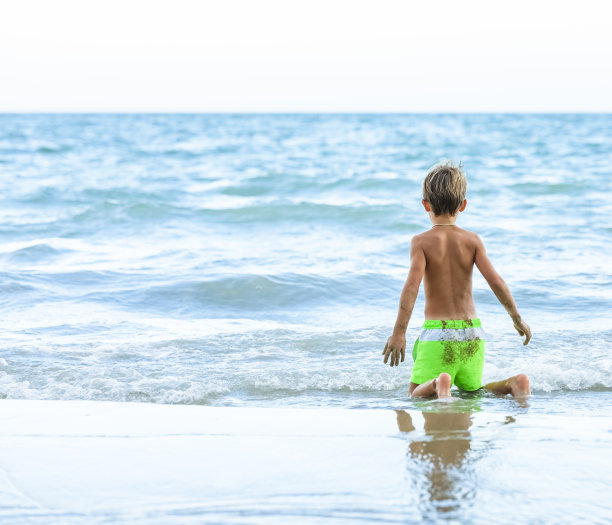 在一个阳光明媚的日子里，那个穿着游泳衣的孩子在沙滩上玩耍。背景是海滩和海浪。图片下载
