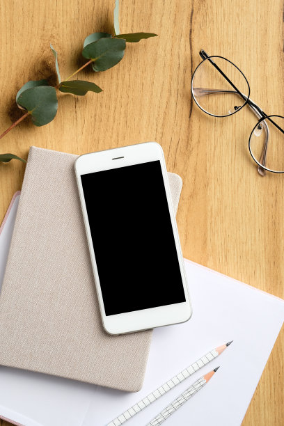 手机放在木质办公桌边，桌子上有女性眼镜，纸笔记本，记事本，桉树叶。平铺，俯视图，拷贝空间。图片下载