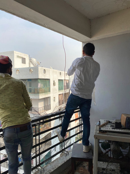 在印度加济阿巴德的住宅阳台上，两个认不出来的亚洲人正在安装防鸽子网，背景是城市扩张的城市景观图片下载