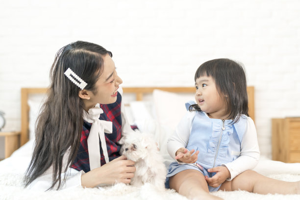 两个年轻的亚洲女人和一只狗在床上。白色的马耳他小狗和一个女孩躺在床上。图片下载