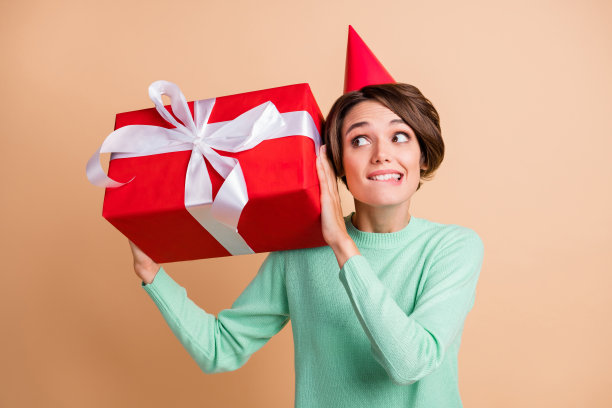 小女孩的照片摇礼物盒好奇感兴趣好奇咬嘴唇戴派对帽孤立在米色背景图片下载