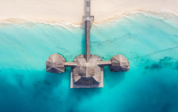 在坦桑尼亚桑给巴尔岛的白色沙滩上，棕榈树茅草屋顶的棚屋被青绿色的印度洋海浪冲刷着。图片下载