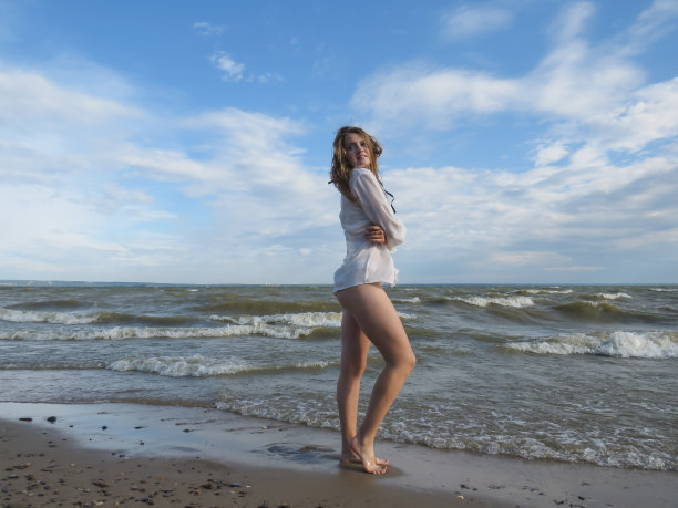 一个年轻的金发女人披头散发，穿着白色的衬衫和比基尼，一双晒黑了的腿在潮湿的沙滩上，背景是海浪和蓝色多云的天空，在一个炎热多风的夏天图片下载