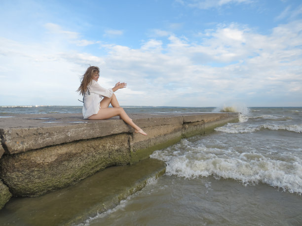 年轻的金发女郎穿着白色的衬衫和比基尼，晒黑了的腿在海滩上，背景是海湾的波浪和多云的天空享受新鲜的海风和丰富多彩的风景图片下载