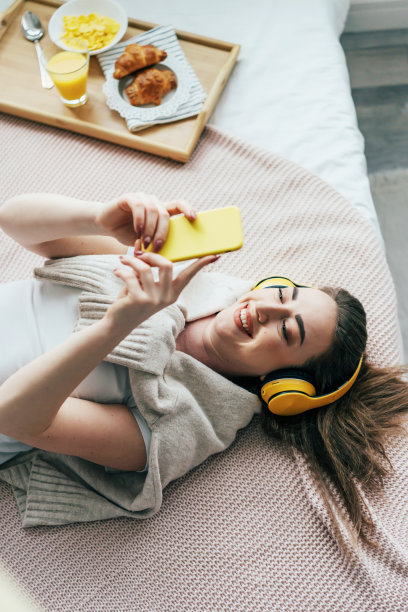 一位年轻迷人的女学生戴着黄色耳机，躺在床上一边打电话一边听音乐。图片下载