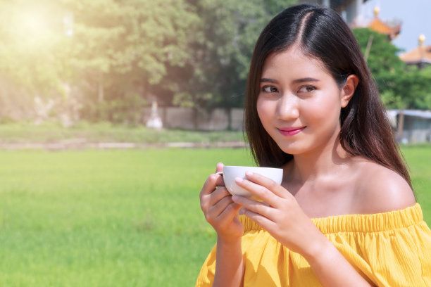 快乐的微笑年轻的妇女穿着黄色的裙子拿着白色的杯咖啡或茶看着稻田里的东西。概念的生活方式放松图片下载