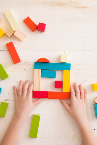俯视图上的孩子的手玩彩色的木砖在白色的桌子背景。男孩建筑与木制构造器。教育的概念图片下载