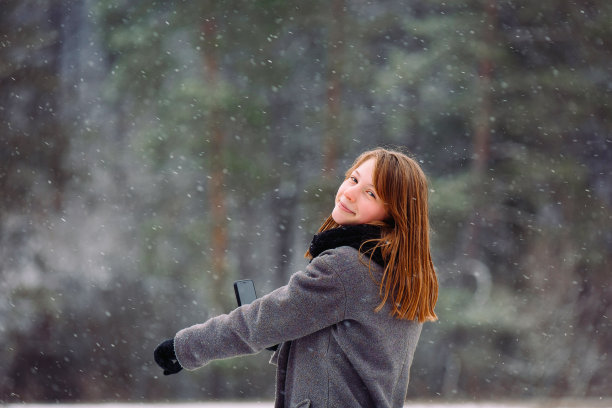 一个白人女孩在冬天的森林里，看到远处的东西，伸出食指，拿着手机，微笑着看着相机。图片下载