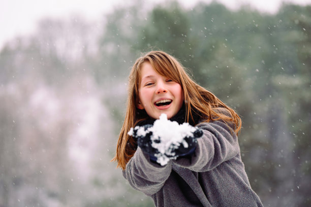 一个女孩面带真诚的微笑，拿着一个雪花球对着镜头。这张照片的背景是冬天被雪覆盖的森林。图片下载