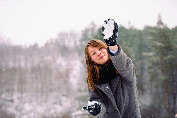 一个漂亮的红鼻子女孩微笑着向镜头扔雪球，背景是冬日的景色。图片下载