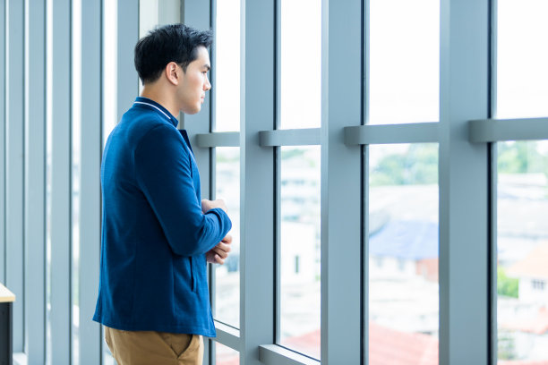 一个开朗成熟的亚洲年轻商人的肖像穿着西装的男人在蓝色夹克和蓝色衬衫看着窗户在办公室的背景。图片下载