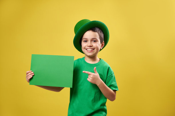 穿着绿色衣服，戴着爱尔兰帽的快乐男孩把他的食指放在一张有拷贝空间的纸上，微笑着在黄色的背景上摆姿势。圣巴特里克节图片下载