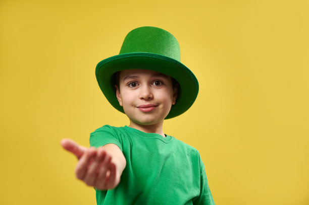 戴着爱尔兰绿色帽子的小男孩伸出手掌对着相机。圣帕特里克节的概念。复制空间和黄色背景图片下载