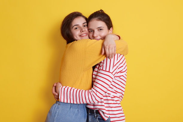 两个欧洲年轻美丽的黑发女孩隔着黄色的背景互相拥抱，微笑着看着镜头，穿着休闲的服装，互相表达爱意，思念，一直没有见过对方。图片下载