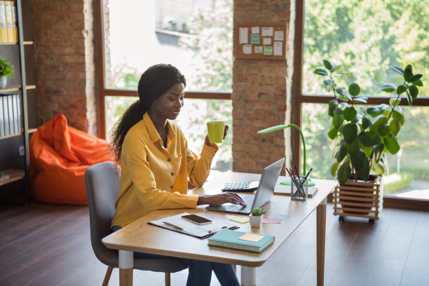 照片的乐观卷曲的黑发漂亮的女孩喝茶型笔记本电脑坐在桌子旁穿黄色衬衫在家图片下载
