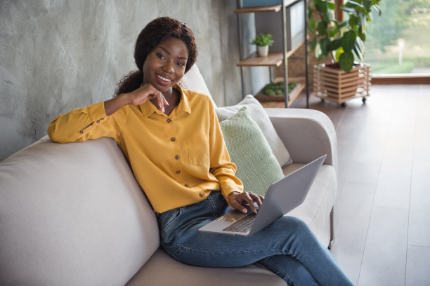 照片肖像的非洲裔美国女孩与笔记本电脑坐在沙发上微笑在现代办公室室内图片下载