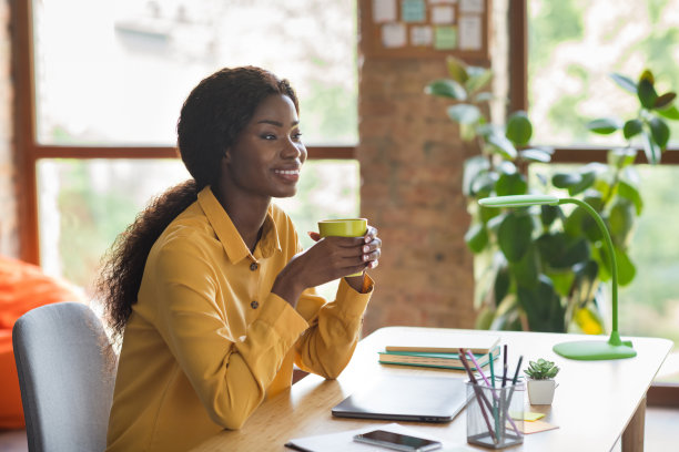 快乐的非洲裔美国女商人的侧面照片，看坐在桌子上，在办公室工作室内喝咖啡休息图片下载