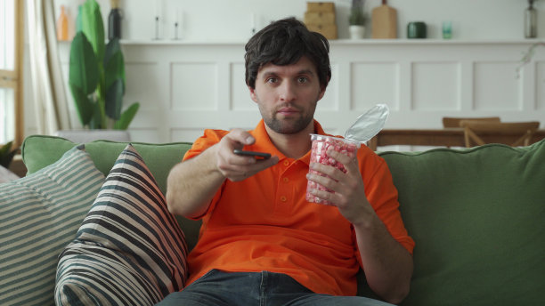 男人坐在沙发上，吃着爆米花，拿着遥控器切换频道图片下载