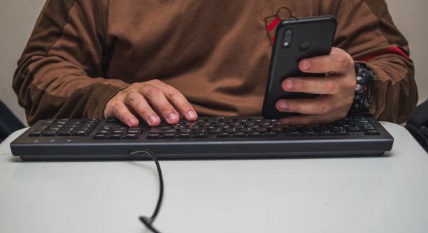 特写手放在黑色的电脑键盘上，拿着手机，放在白色的桌子上图片下载
