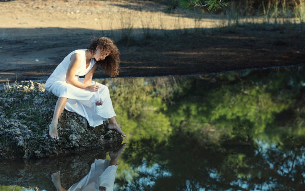 一位穿着白色连衣裙、一头卷发的年轻女子伤心地坐在湖边。图片下载