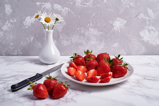 成熟的有机草莓在白色陶瓷盘子和洋甘菊花花瓶在灰色大理石背景，复制空间。健康食品理念，静物生活图片下载