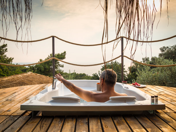 一个英俊的成年男子躺在一个豪华的异国情调温泉度假酒店阳台上的热水浴缸与日落的海景。图片下载