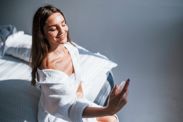 使用香水。房间里的床上坐着一个穿着白色衣服的漂亮的黑发女人的画像。漂亮的灯光图片下载