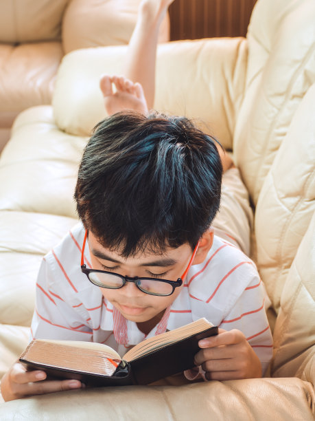 可爱的小孩子穿着休闲的衣服，微笑着躺在沙发上看书，学习，快乐地学习。图片下载