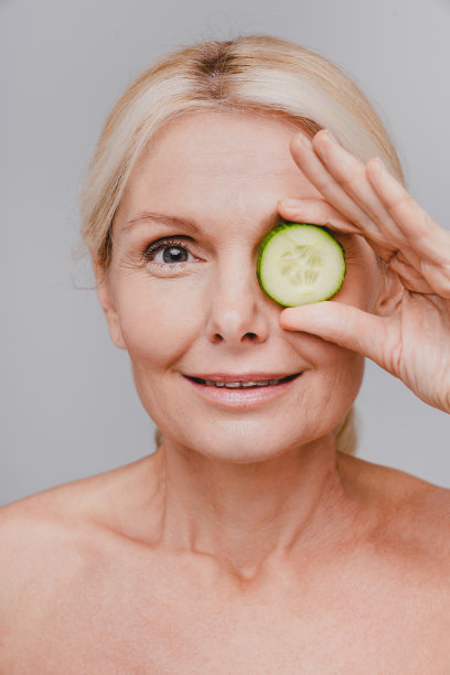 垂直裁剪的肖像中年迷人的女人拿黄瓜片在眼睛孤立在灰色的背景图片下载